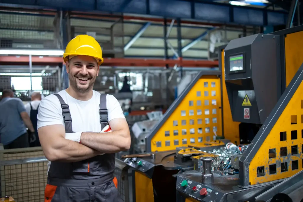 Homem operador de máquinas em fábrica, usando equipamento de segurança, com sorriso confiante, destacando a importância de treinamentos obrigatórios para segurança e eficiência