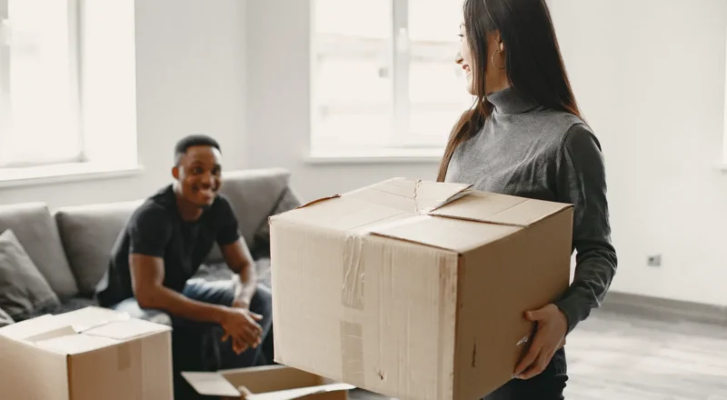 Pessoa em um apartamento organizado com caixas de mudança empilhadas, enquanto um ajudante aguarda ao fundo no ambiente, ideal para ilustrar um checklist de mudança residencial sem estresse antes do grande dia.