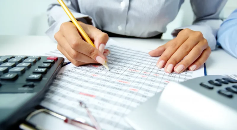 Reforma Tributária: O Que Muda na Prática para Pequenas e Médias Empresas: Analista revisando uma tabela financeira em uma folha de planilha, com calculadora ao lado e lápis apoiado sobre os dados. Foto de escritório que remete a planejamento e impostos para empresas.