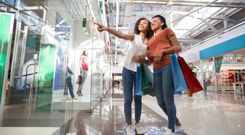 Duas pessoas caminhando em um shopping, com sacolas de compras nas mãos, cenário que representa Ativações Físicas em Shopping Centers: Por Que o Presencial Voltou a iImportar no Marketing e o retorno das experiências presenciais.