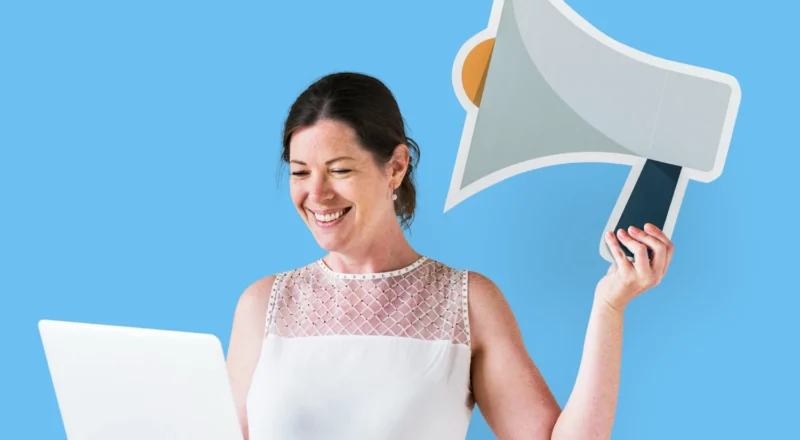 Retenção como Nova Métrica de Sucesso: o Que Times de Growth Precisam Aprender com o Mercado de Assinaturas: Mulher sorridente em fundo azul segurando um notebook e outro dispositivo tipo megafone, sugerindo comunicação e marketing, com foco em retenção e sucesso em assinaturas.
