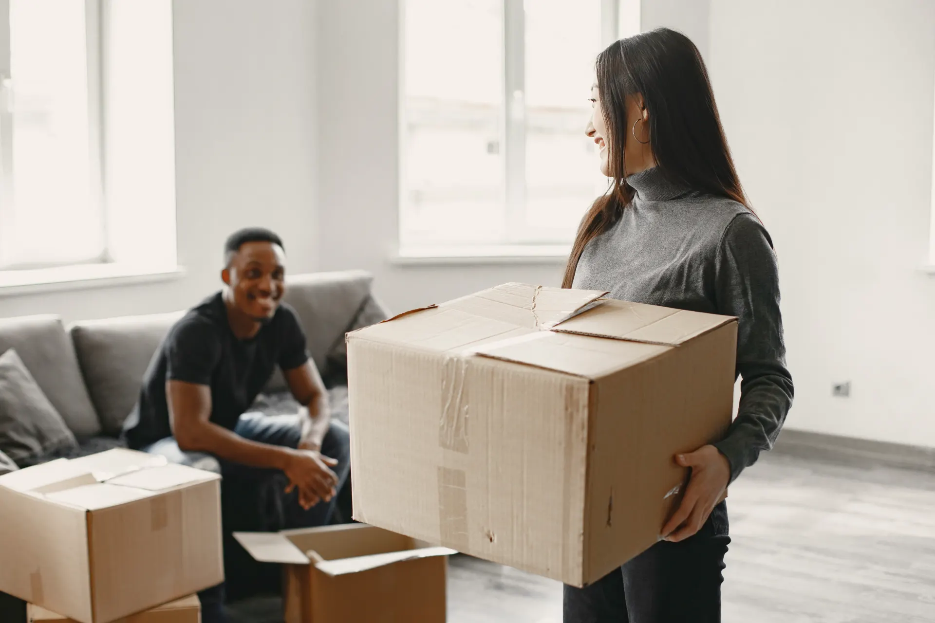 Pessoa em um apartamento organizado com caixas de mudança empilhadas, enquanto um ajudante aguarda ao fundo no ambiente, ideal para ilustrar um checklist de mudança residencial sem estresse antes do grande dia.
